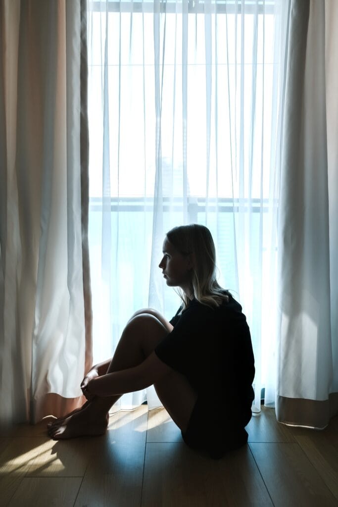 Silhouette of a person sitting on the floor in front of a large window with sheer curtains, daytime. They are sitting with knees drawn to chest and resting their arms on their legs, looking contemplatively out the window.